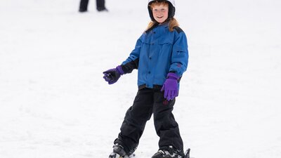 A boy skis on a gentle slope in a snowplough stance. The background is covered in snow.